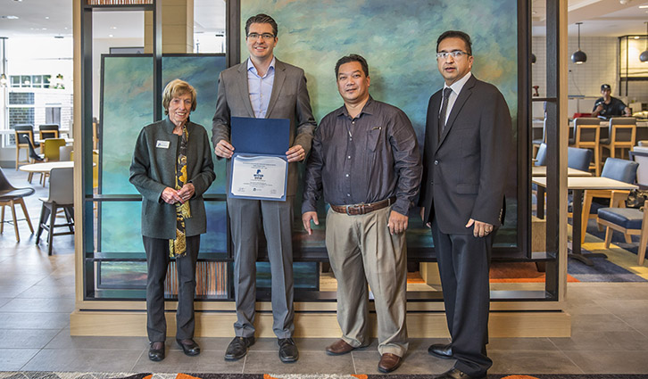 Four professionals indoors posing for an award ceremony, with a framed certificate held by the man second from the left.
