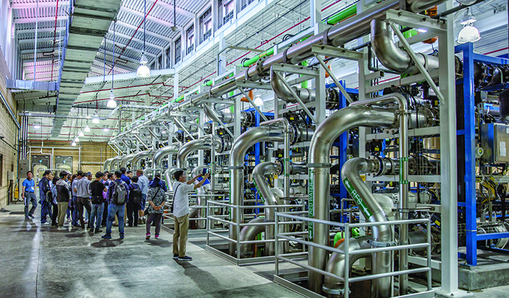 Group of visitors on a guided tour inside an industrial plant with large stainless steel piping and processing equipment.