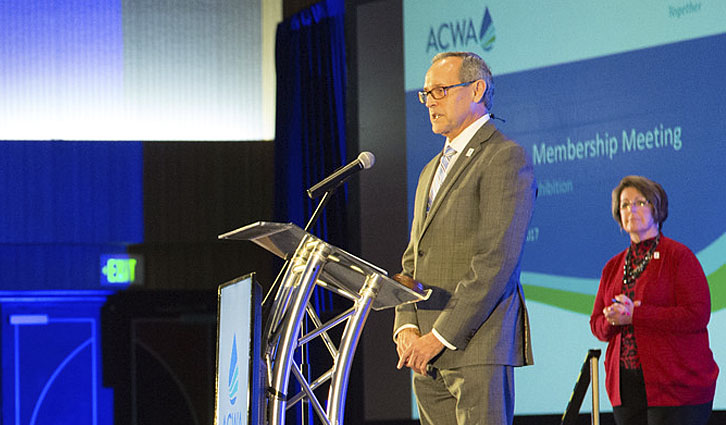 Man in a gray suit speaks at a podium on a stage during a membership meeting; a woman in red stands behind him.