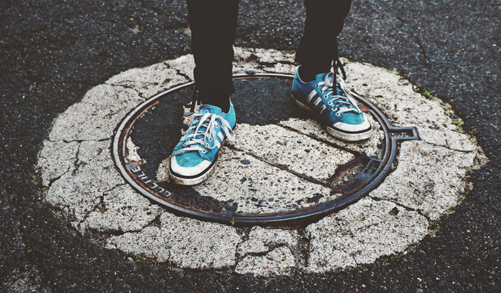 Person wearing blue sneakers stands on a cracked circular manhole cover in an urban street.