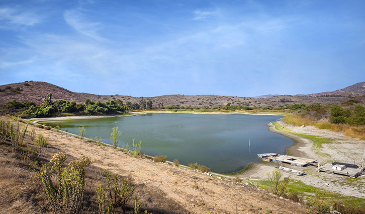Arid landscape with a calm blue-green lake, sandy shores, and distant hills under a clear blue sky.