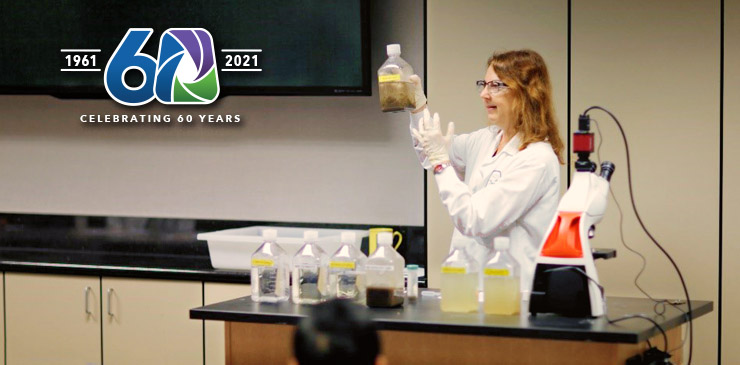 Scientist in a lab coat and gloves holds a bottle of liquid at a bench with chemical bottles and lab equipment.