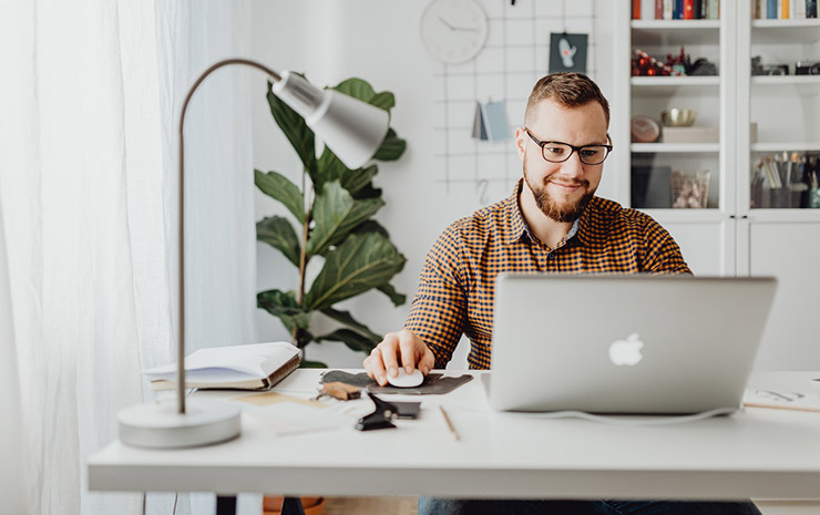 Bearded man with glasses works on a MacBook at a white desk in a bright home office with plants.