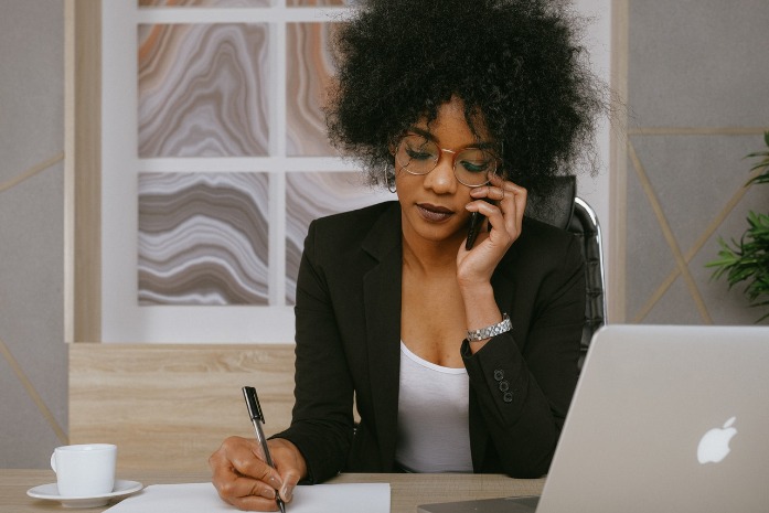 Professional woman with curly hair wearing glasses, in an office, talking on a phone while taking notes.