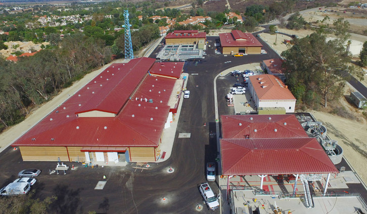 Aerial view of a red-roofed facility complex with roads, vehicles, and surrounding trees.