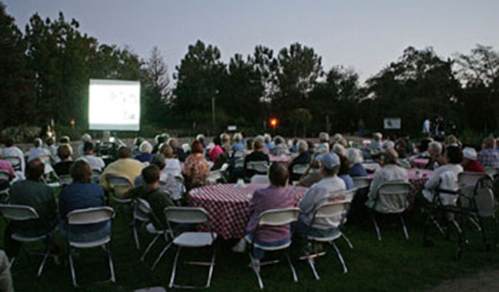 Outdoor community movie night with a large crowd seated on lawn chairs facing a projection screen at dusk.