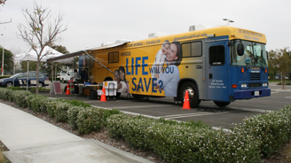 Blue and yellow mobile health bus with a 'Life Will You Save' ad parked in a parking lot beside a pop-up tent.