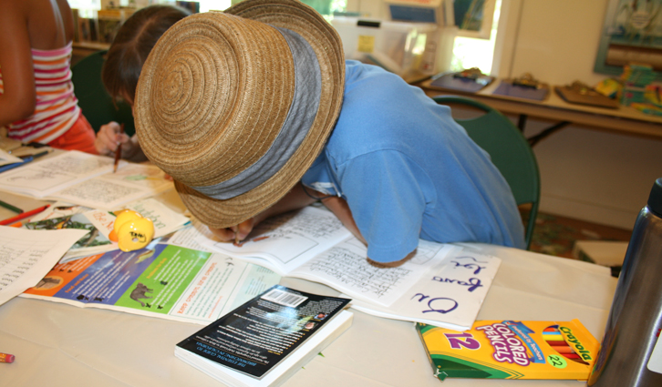 Child wearing a straw hat leans over a table of worksheets and coloring supplies, focused on a task.