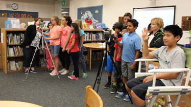 Diverse group of students lined up in a school library as a camera on a tripod records their project.