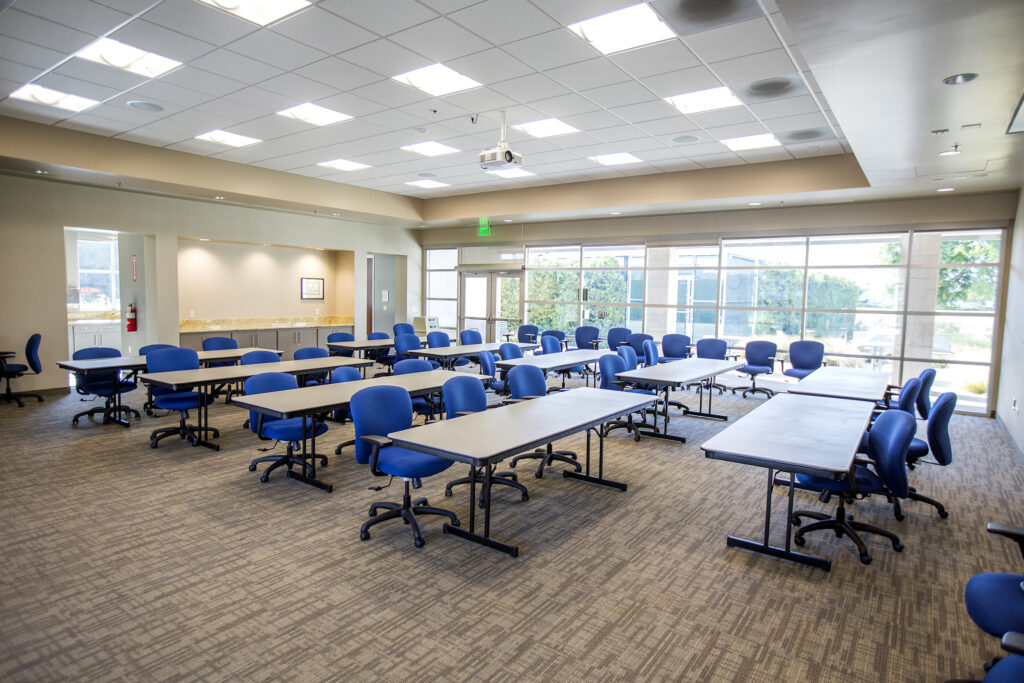 Training room with blue chairs and long tables arranged in rows, projector ceiling-mounted, and large windows.