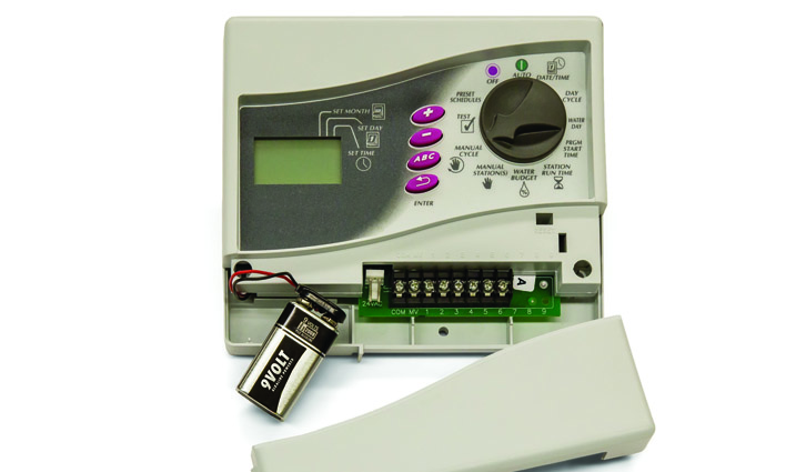 Open home security alarm control panel showing wiring terminals, a 9V battery, and the detached front cover.
