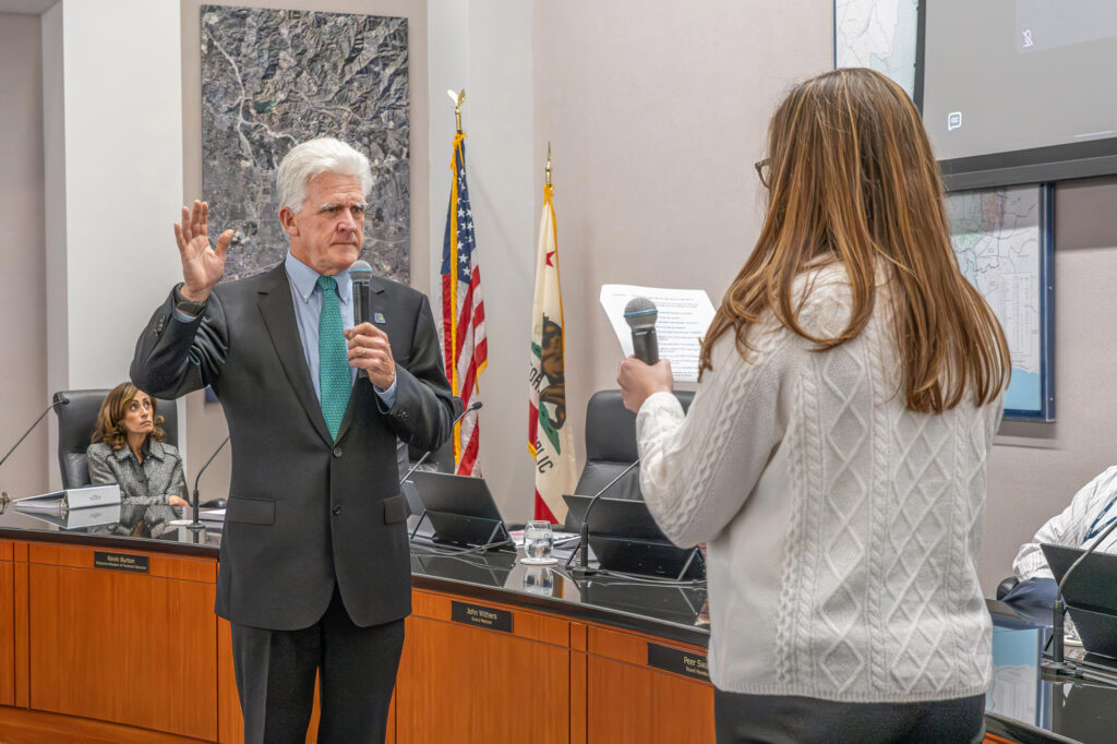 Public official addresses attendees in a boardroom, holding a microphone as a journalist asks questions.