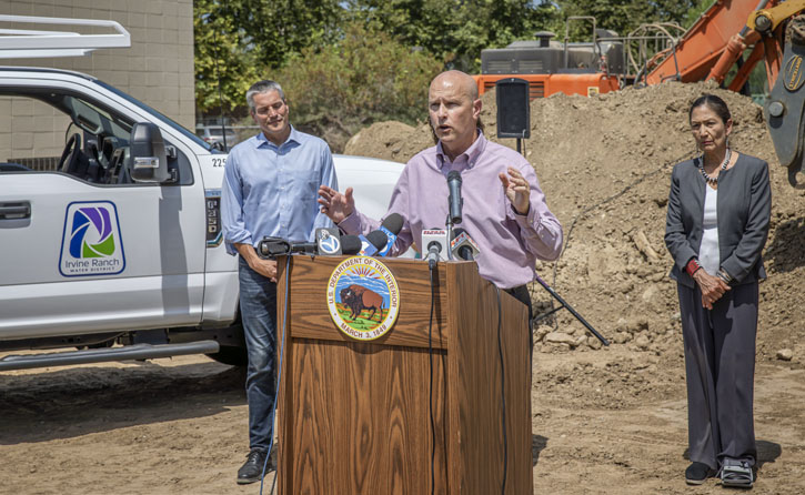 Bald man speaks at a podium with microphones at a construction site, flanked by two officials.
