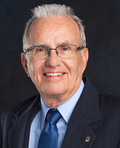 Older man with silver hair and glasses in a navy pinstripe suit, light blue shirt, and blue tie, smiling.