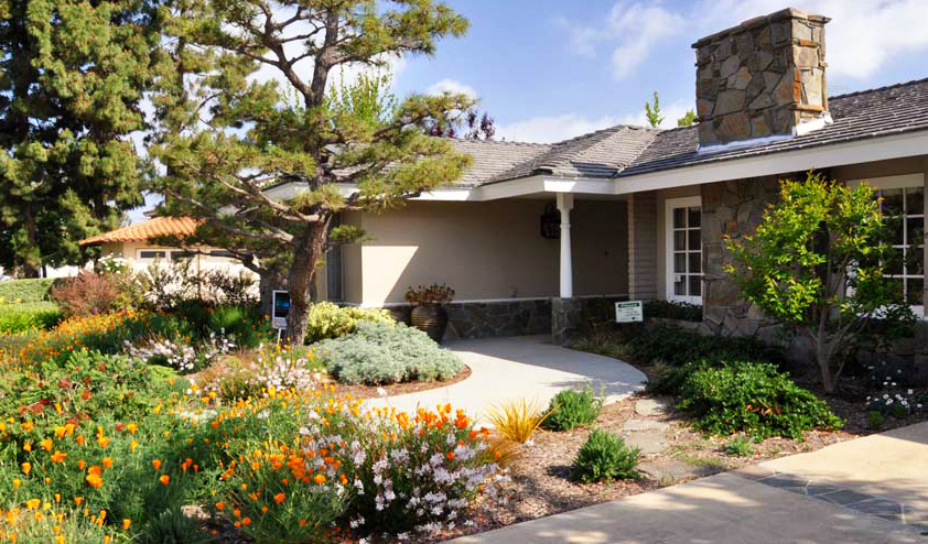 Residential home with a stone chimney, landscaped garden and colorful flower beds along a curved walkway.