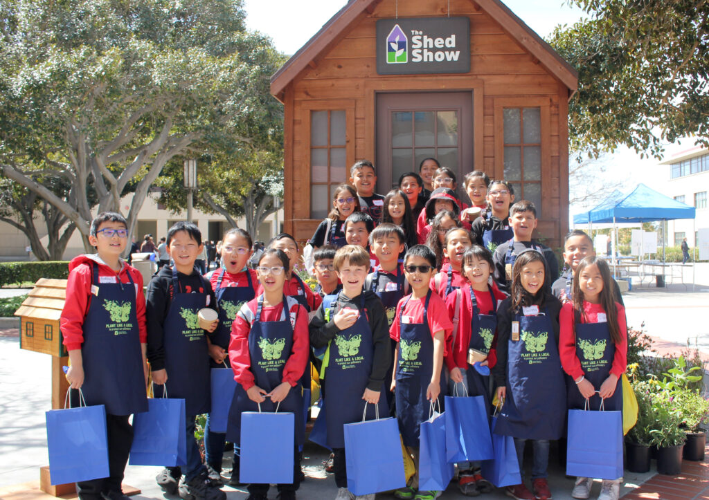 Group of smiling children in red shirts and blue aprons posing in front of a wooden shed with The Shed Show sign.