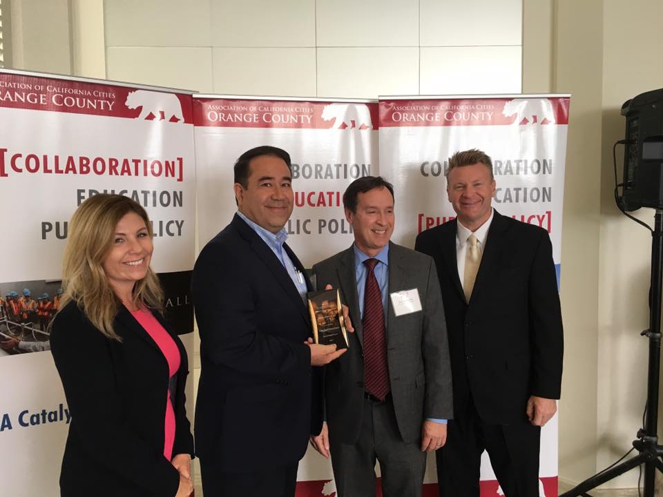 Four professionals at an Orange County event, one man receiving a trophy from another with banners in the background