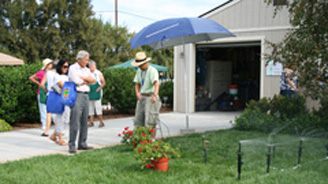 Group of neighbors gathered on a driveway beneath a blue umbrella in a suburban yard.
