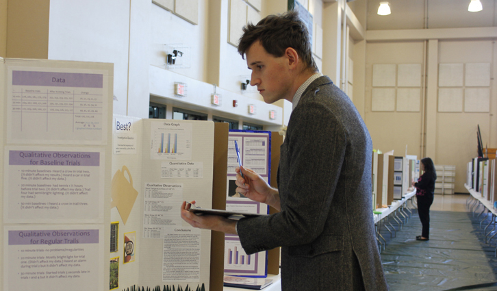 Man in a gray blazer examines a research poster with charts at a university poster session in a large hall.