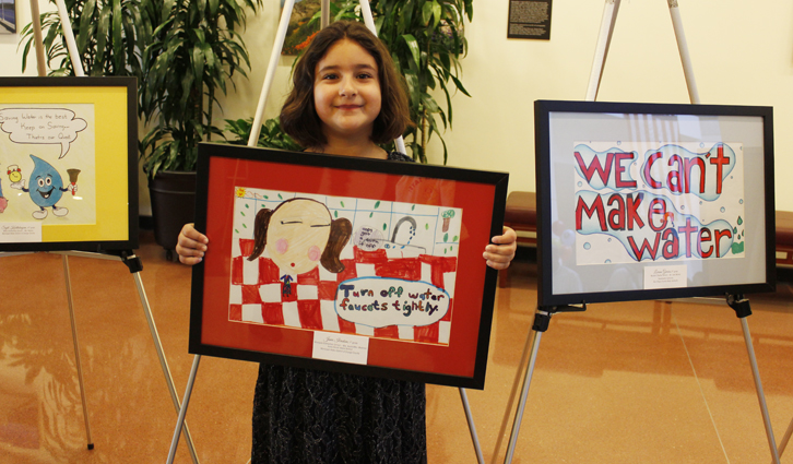 Young girl holding a framed artwork about turning off water faucets, displayed with two other framed pieces.