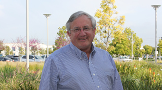 Smiling older man in a light blue striped shirt outdoors with greenery and trees in the background.