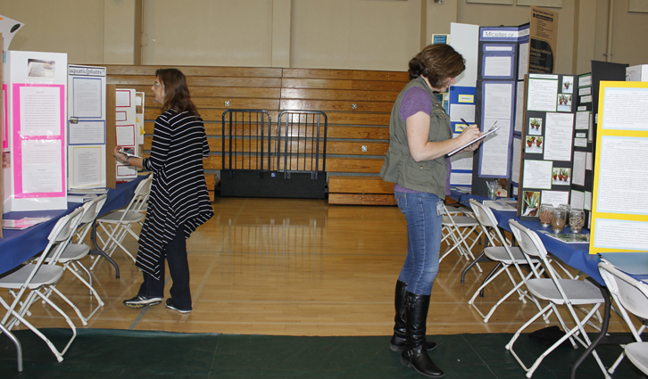 Two women examine science fair posters set up on folding tables in a gymnasium