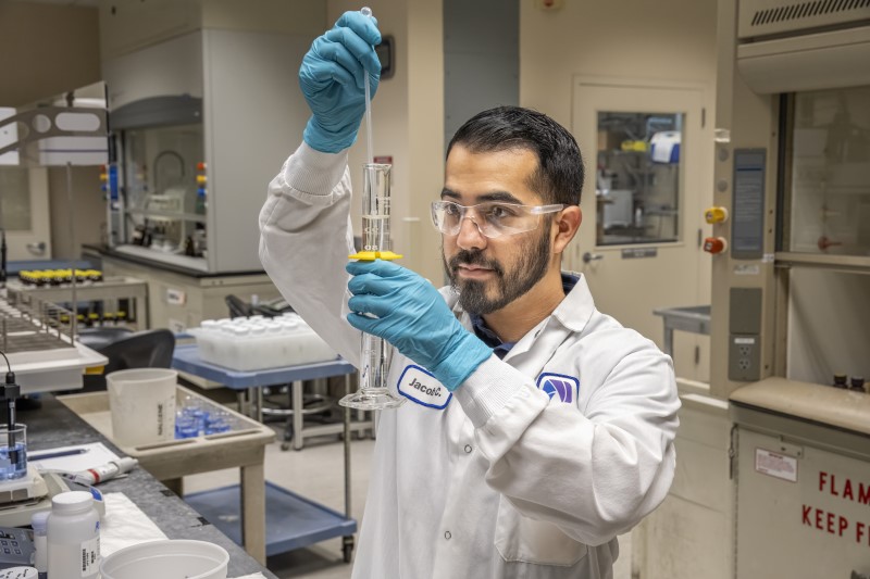 Lab technician in safety goggles and blue gloves pours liquid from a burette into a graduated cylinder in a laboratory.