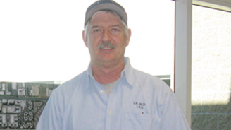 Male technician in a light blue work shirt and cap standing indoors beside electronic equipment.