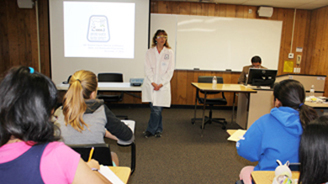 Instructor in a white lab coat lectures at the front of a classroom to students, with a projector screen and whiteboard behind.