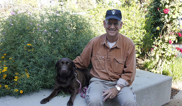 Older man in an orange shirt sits on a concrete bench with a chocolate Labrador in a sunny garden.