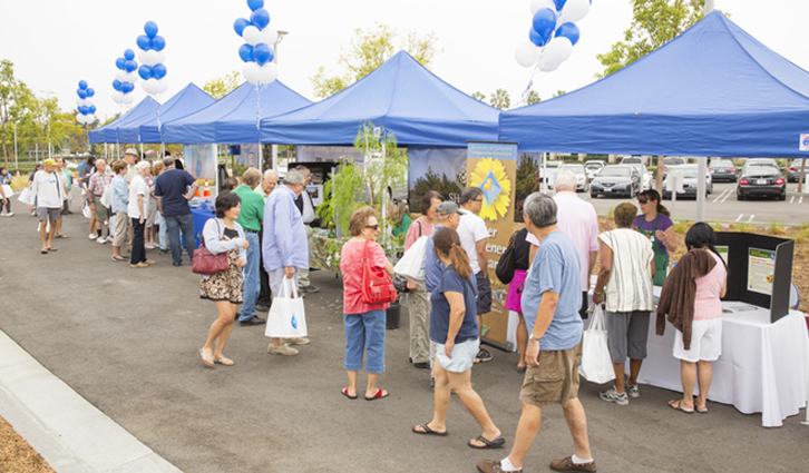 People queue at an outdoor community fair under blue canopies with balloons, visiting vendor booths and tables.