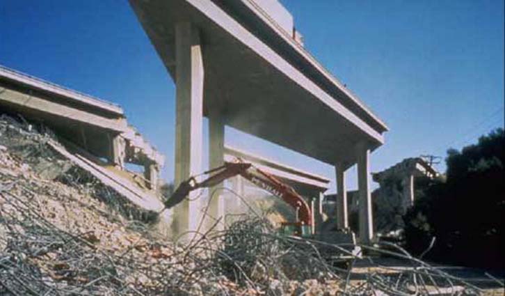 Demolition site showing a partially collapsed concrete overpass, rubble, and an excavator at work.