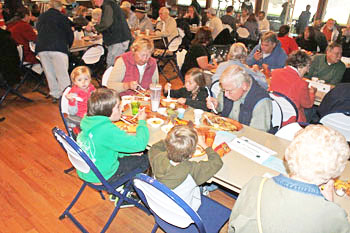 Crowded hall with seniors and children seated at long tables, sharing a meal and chatting.