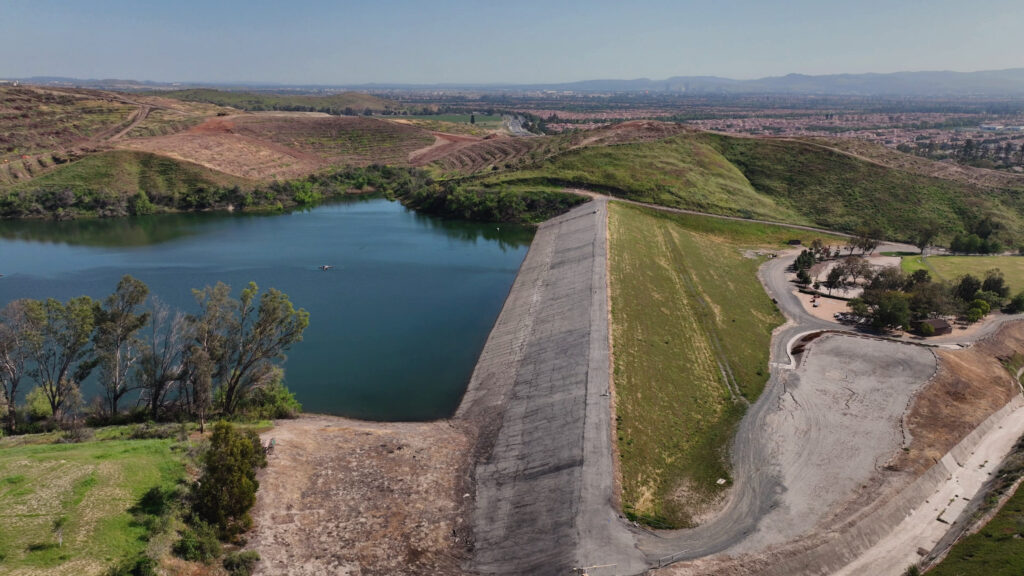 Aerial view of a reservoir with a concrete dam wall, green hills, and a distant city on the horizon.