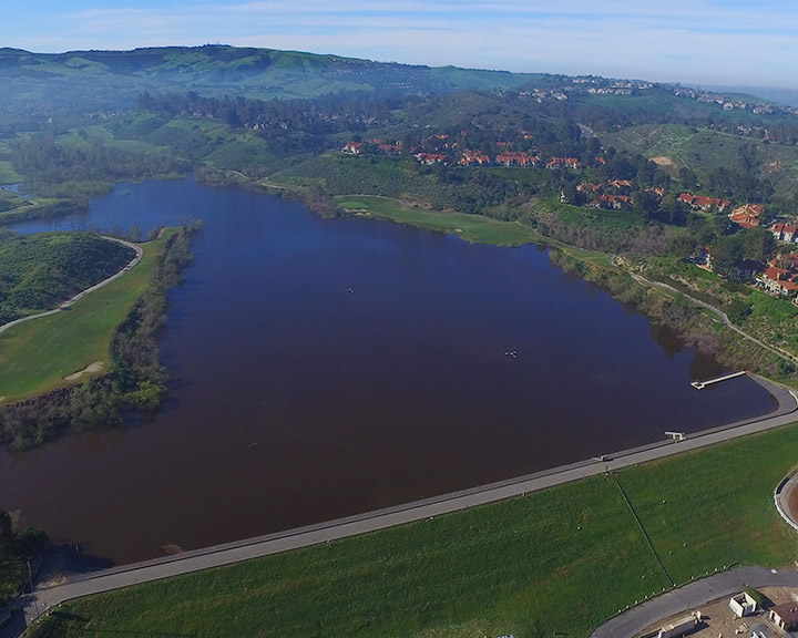 Aerial view of a large lake bordered by green hills and residential neighborhoods, with a road along the shoreline.