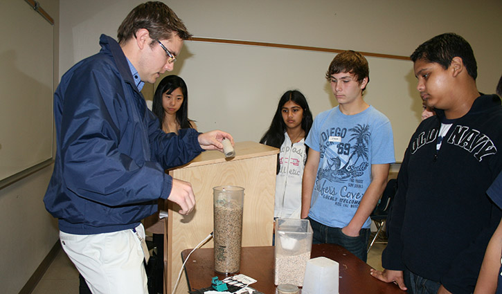 Instructor demonstrates a science experiment to students, pouring a substance into a tall column while observers look on.