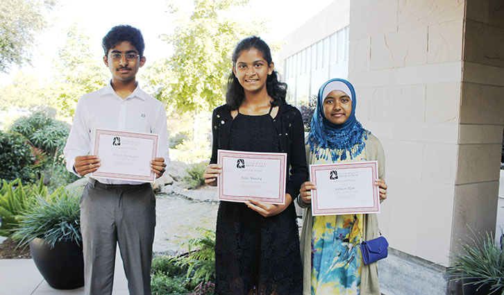 Three students stand outside a campus building, each holding a certificate and smiling at the camera.