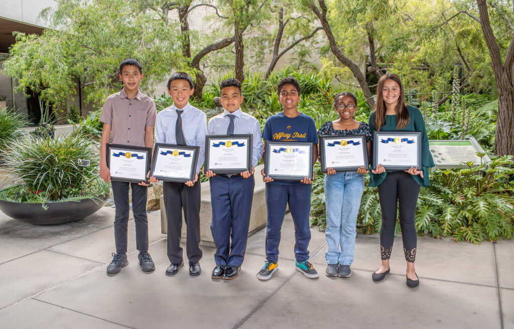 Six diverse students stand outdoors holding award certificates.