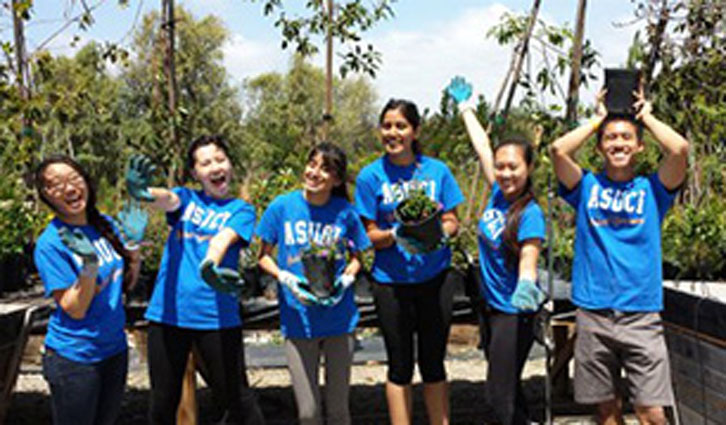Group of volunteers in blue shirts wearing gloves, holding saplings outdoors and smiling.