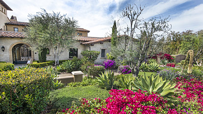 Mediterranean-style villa courtyard with stone walls, flowering shrubs, and vibrant cacti under a sunny sky.