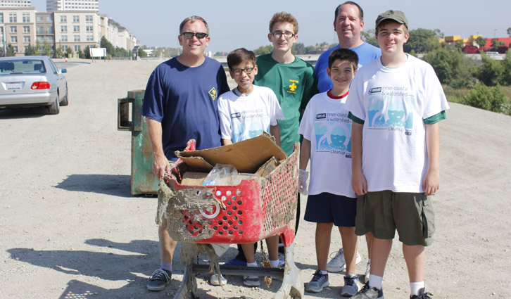 Six volunteers pose with a red shopping cart filled with debris during an outdoor cleanup on a sunny day.