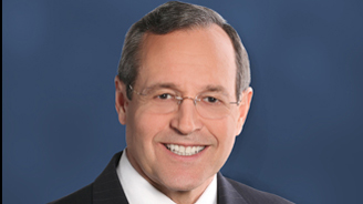 Professional headshot of a middle-aged man in a dark suit and white shirt, smiling, against a blue background.