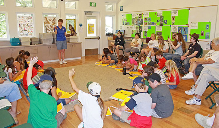 Instructor leads a classroom workshop as children sit in a circle on the floor, many raising their hands.