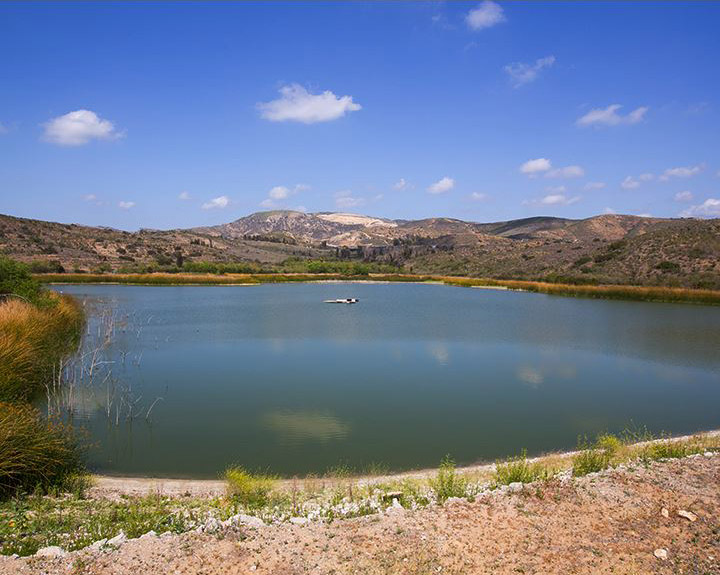 Calm, circular lake bordered by rocky shore and dry hills under a bright blue sky with scattered clouds.