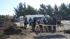 People gathered around an outdoor excavation site with a dirt pile, wooden barrier, and a white van in the background.