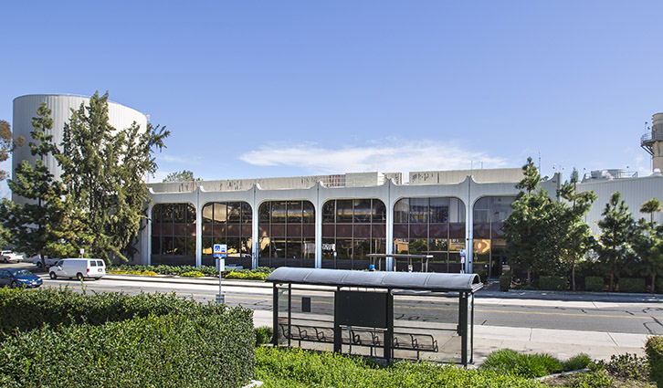 Bus stop in front of a mid-century modern building with arched windows, trees, and a blue sky.