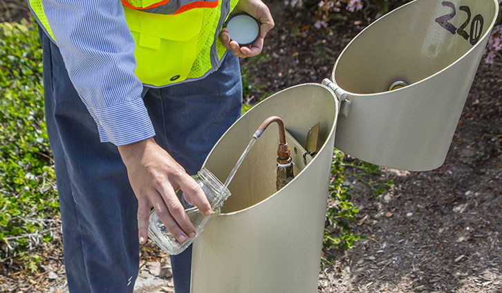 Worker in a high-visibility vest collects a water sample using a dual-compartment field sampler.