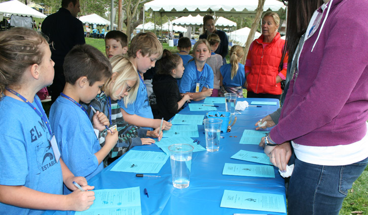 Children signing forms at a long blue table outdoors, supervised by adults at a park event.