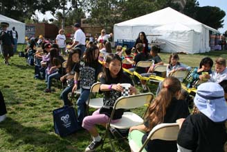 Children and families sit on folding chairs at an outdoor community event on a grassy field beside white tents.
