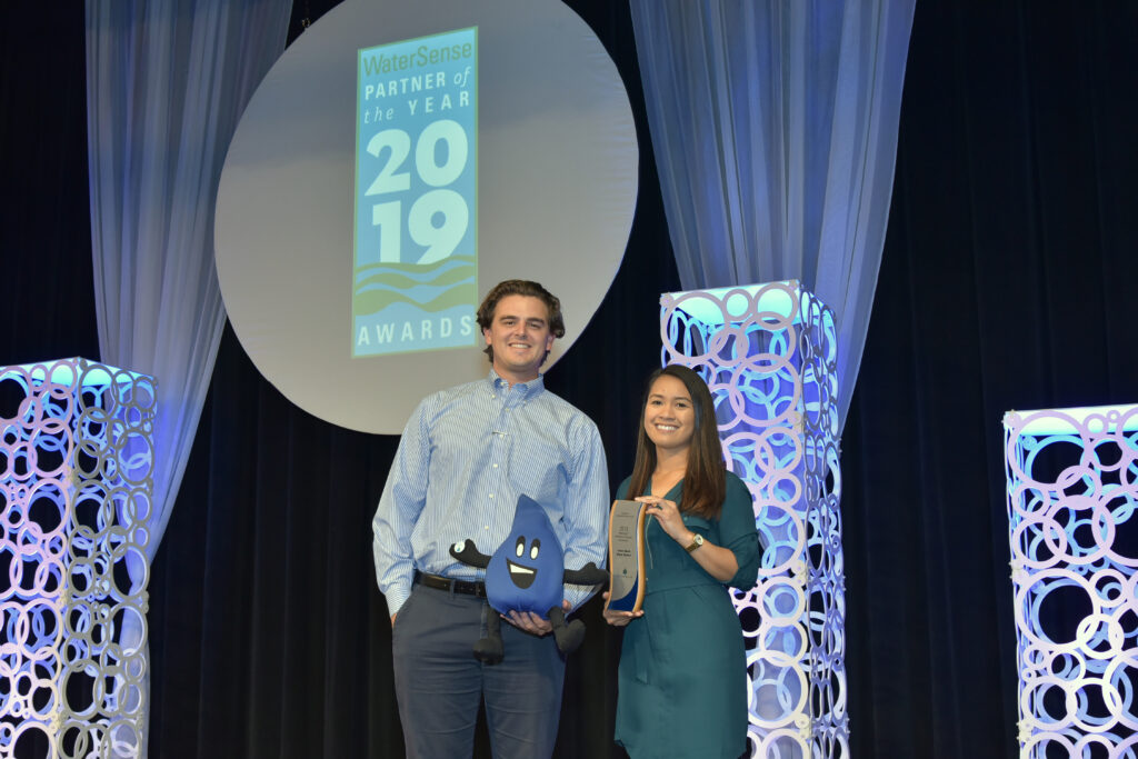 Two people on stage at the WaterSense Awards 2019, man with a blue droplet plush and woman with a plaque, backdrop.